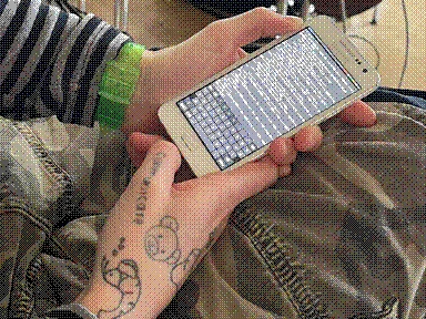 Two hands are holding a Samsung Galaxy A3, with the screen displaying the terminal. One of the hands has a tattoo on the thumb that says 'communicare', as well as a shrimp tattoo.