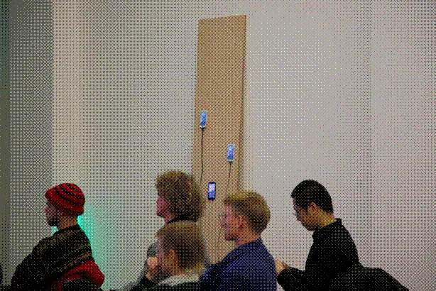 A crowd listening to a presentation by Aymeric Mansoux. In the background is an installation of 3 phones hosting web-servers.
