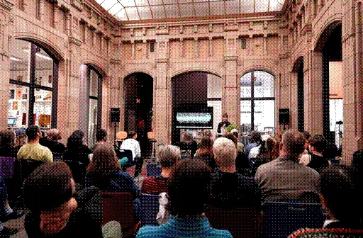 A photo of the symposium with Dasha Ilina presenting her work, with the backs of the listening crowd.